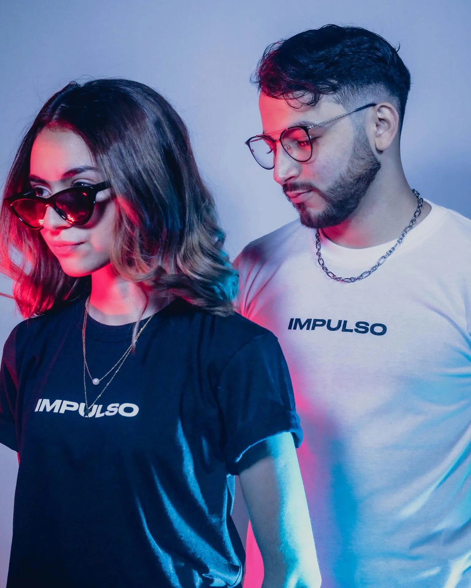 Two young adults, a man and a woman, standing side by side wearing black and white branded t-shirts with the word 'IMPULSO'.